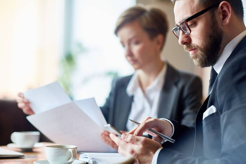 Businessman messaging in smartphone while his colleague looking through papers